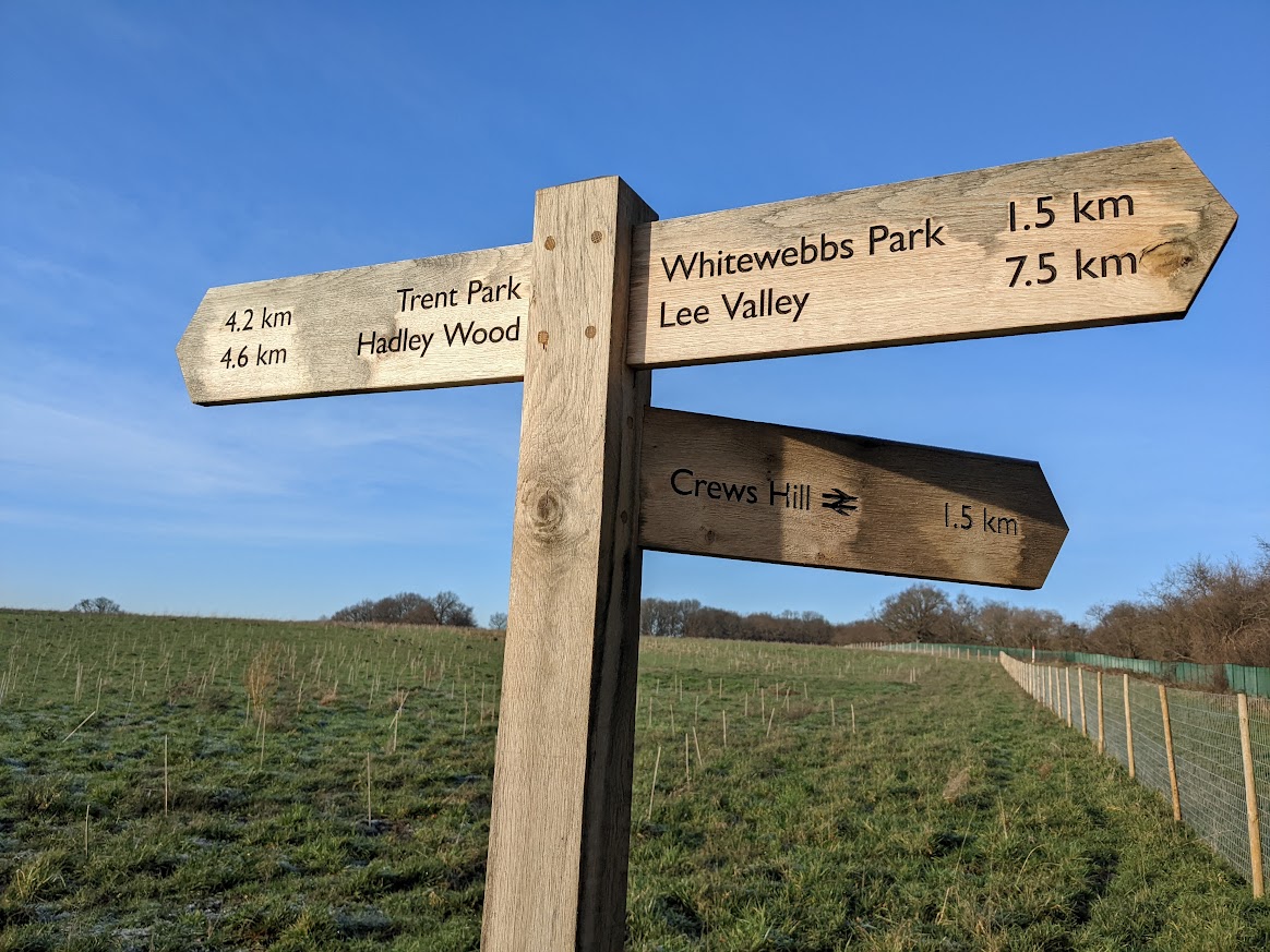 Wooden footpath sign pointing to Crews Hill, Whitewebbs Park, Trent Park and Lee Valley, with open greenfield land behind it.